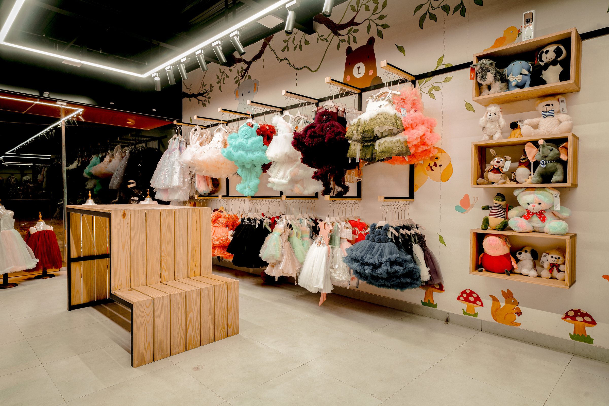 Children's dresses and plush toys arranged around a wood display beneath a painted woodland mural