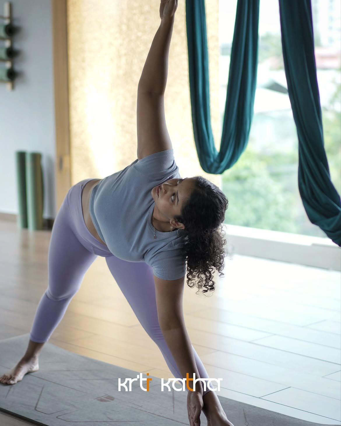 Woman holding a wide side-bend pose beside teal aerial silks in a bright studio